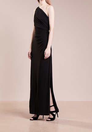 Femme portant une longue robe noire asymétrique avec une fente latérale et des sandales noires à talons hauts, debout devant un fond uni.