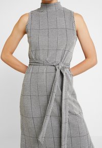 Vestido sin mangas a cuadros en blanco y negro, con un cuello alto, cinturon en la cintura y una tela texturizada con un patrón de rejilla.