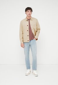 Μπεζ μπουφάν με καφέ κουμπιά, δύο μπροστινές τσέπες, φορεμένο πάνω από μπορντό πουκάμισο και ανοιχτό μπλε τζιν, σε συνδυασμό με λευκά και μπεζ αθλητικά παπούτσια.