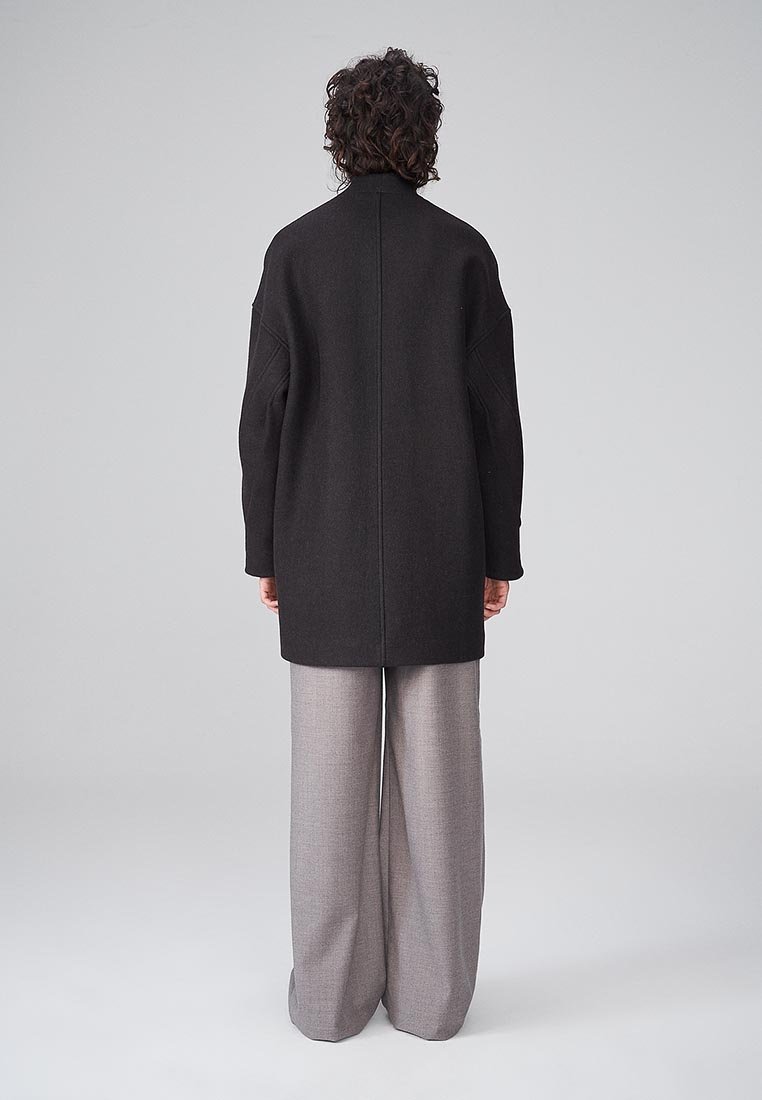 Lungo cappotto di lana nera con colletto alto e vestibilità ampia, abbinato a pantaloni grigi larghi, mostrato di spalle su uno sfondo neutro.