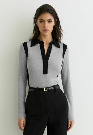 Žena stojící u bílé zdi, má na sobě šedý dlouhý rukáv s černým límečkem a černé kalhoty s pásem, dívá se vpřed.