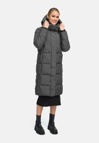 Cappotto lungo imbottito di colore grigio scuro, con colletto alto, chiusura con zip e cappuccio oversize. Abbinato a una gonna nera a coste e stivali neri.