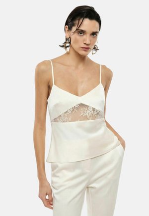 Donna con capelli corti e scuri che indossa grandi orecchini geometrici, una canottiera in raso bianco con pannello in pizzo e pantaloni in raso bianchi abbinati.