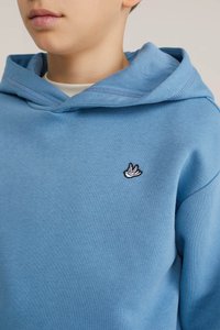 Copil care poartă o hanorac albastru cu un mic emblema albă în formă de pasăre pe piept, arătând gâtul, partea inferioară a feței și partea superioară a trunchiului.