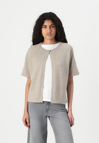 Beige strikket cardigan med korte ermer, rund halslinje og en enkelt knappåpning foran, båret over en hvit skjorte og grå jeans.