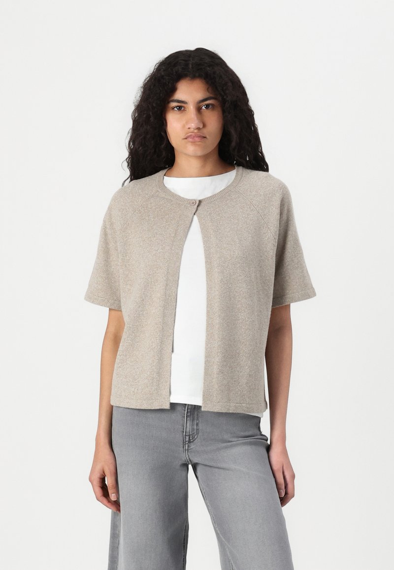 Beige strikket cardigan med korte ermer, rund halslinje og en enkelt knappåpning foran, båret over en hvit skjorte og grå jeans.