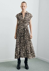 Maxi-jurk van donkerbruine stof met crème abstracte luipaardvlekken, korte mouwen, V-hals, getailleerde taille en een vloeiende rok, gecombineerd met zwarte laarzen.