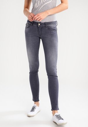 Person iført tætsiddende mørkegrå jeans, grå t-shirt og hvide sneakers stående på en lys baggrund med hænderne foldet.