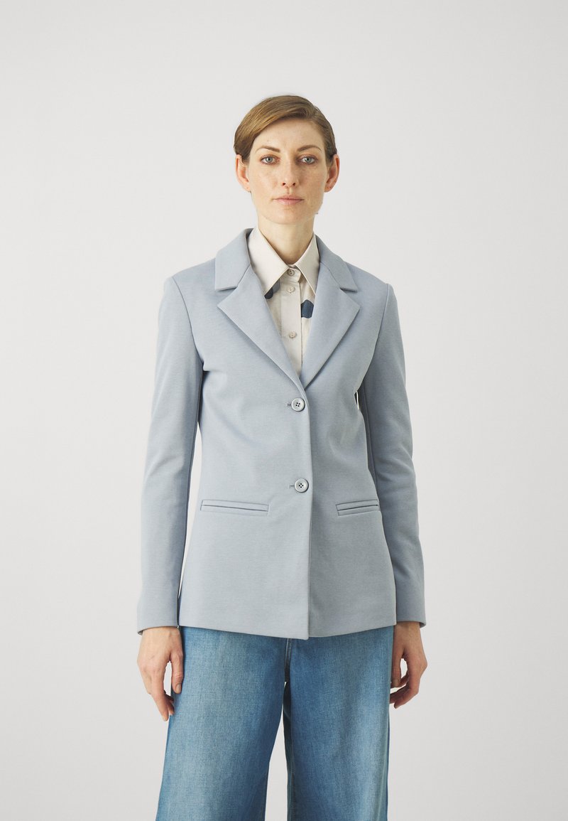 Lichtblauw op maat gemaakt blazer met twee knopen aan de voorkant, scherpe kraag en twee zijzakken, gedragen over een patroonshirt en wijde jeans.