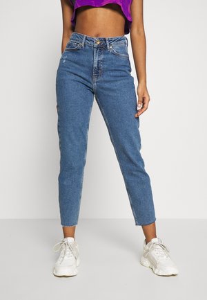 Kobieta ubrana w niebieskie dżinsy z wysokim stanem i surowo wykończonymi nogawkami oraz białe sneakersy, stojąca na tle jednolitego białego tła.