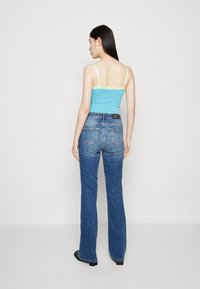 Canotta azzurro chiaro con rifiniture in pizzo, abbinata a jeans a vita alta blu scuro svasati. Modella mostrata di spalle, scarpe nere visibili.