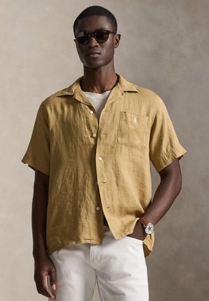 Hombre con gafas de sol oscuras, camisa mostaza de manga corta abotonada sobre pantalones blancos, con una mano en el bolsillo y un reloj en la muñeca.