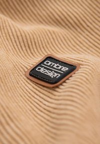 Tessuto a righe beige con una toppa in pelle nera e marrone con l'etichetta "Ombre Design", caratterizzata da lettere in rilievo e texture lisce.