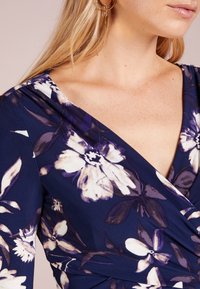 Marinblå klänning med vitt och ljuslila blommönster, v-ringning, mjukt tyg och draperade detaljer vid midjan.