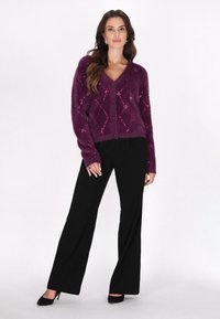 Cárdigan de felpa morado con un escote en V pronunciado, cierre de botones y un patrón de diamantes con acentos en rosa, combinado con pantalones anchos negros y tacones.