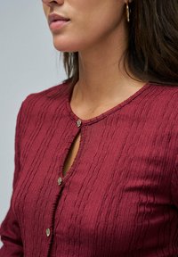 Top de textura burdeos con escote redondo, cierre de botón y un pequeño detalle en forma de lágrima. La tela tiene un patrón de canalé.