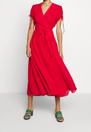 Femme portant une robe portefeuille rouge fluide avec des manches courtes nouées et des sandales à talons rayées vertes, debout devant un fond blanc.