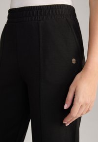 Zwarte tapered sweatpants met een elastische tailleband, voorzien van zijzakken en een klein metalen logo-accent nabij de zak. Gladde textuur.