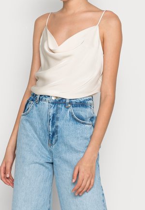 Femme portant un top crème sans manches drapé associé à un jean denim bleu clair taille haute sur un fond uni.
