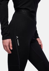 Leggings noirs en tissu extensible, avec une taille haute, une texture lisse et un logo blanc sur la cuisse gauche. Des détails de couture sont visibles.