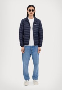 Doudoune navy avec fermeture éclair à l'avant, capuche et design matelassé ; associée à un t-shirt blanc, un jean bleu clair et des baskets beiges.