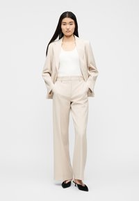 Γυναίκα με μακριά σκούρα μαλλιά, ντυμένη με μπεζ κοστούμι, λευκό αμάνικο τοπ και μαύρες μυτερές γόβες, στέκεται με τα χέρια στις τσέπες σε λευκό φόντο.