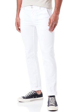 Beine, die weiße, umgekrempelte Hosen tragen, kombiniert mit schwarzen Low-Top-Sneakern mit weißen Schnürsenkeln und Sohlen, stehend auf einer reflektierenden weißen Oberfläche.