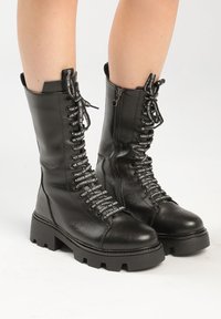 Botas de combate de cuero negras hasta la mitad de la pantorrilla con suelas gruesas tipo lug, cordones negros y blancos con texto repetido, usadas por una persona de pie sobre un suelo blanco.