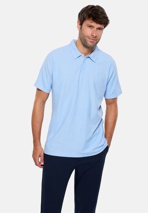 Hombre con camisa polo de manga corta azul claro y pantalones oscuros, de pie con una mano en el bolsillo contra un fondo liso y claro.