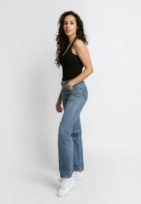 Jeans in denim blu con un design a gamba svasata e tasche frontali, abbinati a un top nero e sneakers bianche, caratterizzati da una vestibilità casual.