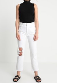 Bílé denimové džíny s obnošenými detaily na kolenou, v kombinaci s černým žebrovaným topem bez rukávů a černými sandály s kulatými kovovými akcenty.