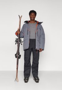 Veste de ski isolante grise avec des accents orange, chemise à manches longues grise, pantalon de ski noir et bottes de ski, tenant des skis avec fixations.