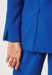 Hand entspannt an der Seite, trägt ein lebhaftes blaues Sakk und passende Hose, mit einem Schlitzdetail an der Ärmelnaht nahe dem Handgelenk.