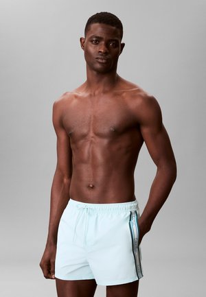 Hemdloser Mann mit dunkler Hautfarbe, der hellblaue Badeshorts mit gestreiftem Calvin Klein-Logo an der Seite trägt und vor einem grauen Hintergrund steht.