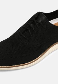 Svart textilskor med ett andningsbart stickat ovandel, snörning och en kontrasterande vit gummisåle med sömdetaljer.