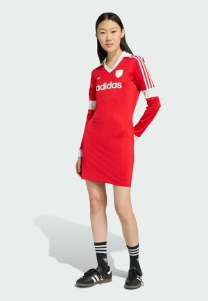 Abito lungo a maniche lunghe rosso con colletto bianco e dettagli bianchi sulle maniche. Logo Adidas ben visibile sulla parte anteriore. Scarpe nere e calze a righe abbinate.