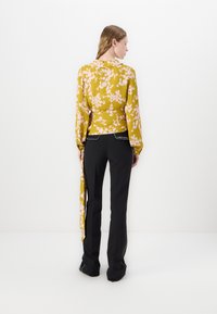 Langärmlige senfgelbe Bluse mit pinkem Blumenmuster, tailliert mit einem Knoten, kombiniert mit schwarzen Hosen mit seitlichem Streifen. Glatte Stoffstruktur.