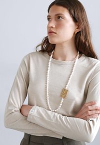 Beige pikk varrukaga topp ümmarguse kaelusega. Omab läikivat kuldset ripatsikaelakee, millel on teerajajate helmed. Tekstuuriga kangas, lõdva lõikega.