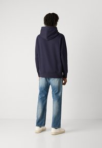 Felpa blu navy con un ampio cappuccio e tasca a marsupio anteriore, abbinata a jeans dritti blu chiaro e sneakers color crema.