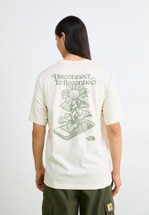Osoba z długimi ciemnymi włosami nosi kremowy t-shirt z zielonym słonecznikiem wyrastającym ze smartfona oraz tekst „Disconnect To Reconnect.”