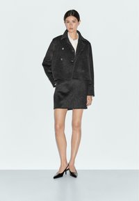 Black cropped wool jacket with a double-breasted design, paired with a matching pencil skirt. Model wears a white shirt and black heels.
