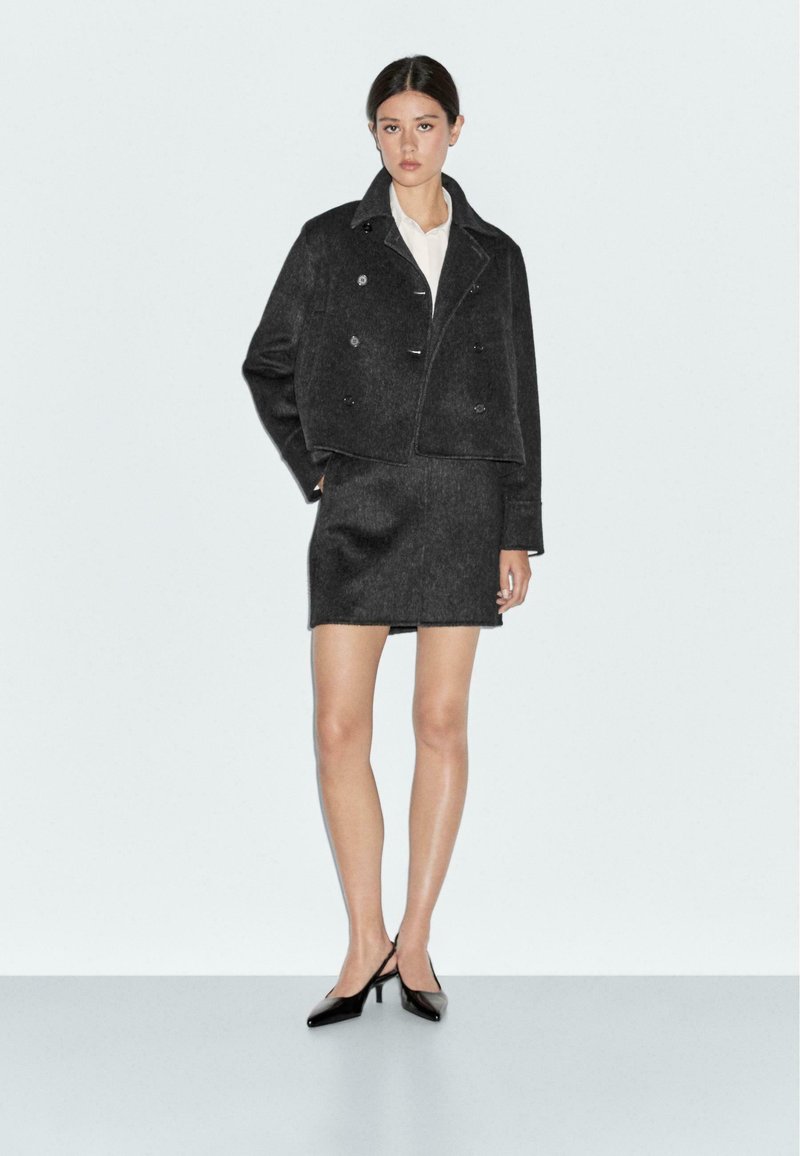 Black cropped wool jacket with a double-breasted design, paired with a matching pencil skirt. Model wears a white shirt and black heels.