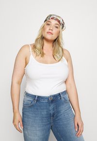 Witte spaghetti top in combinatie met middelblauwe high-waisted jeans. Een bedrukte haarband voegt een kleurrijke accent toe.