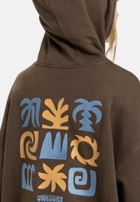 Brauner Kapuzenpullover mit großem grafischem Druck, der auf der Rückseite blaue und orangefarbene abstrakte Muster zeigt. Weiches, strukturiertes Material, lässige Passform.