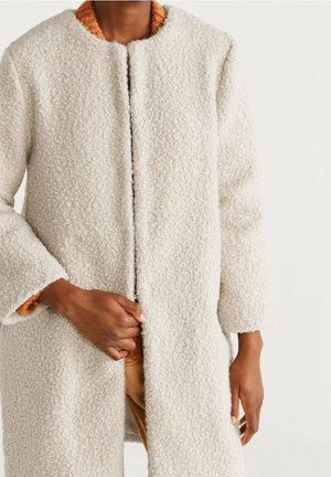 Personne portant un manteau en laine blanc cassé texturé sur un pull couleur rouille, tenant le manteau fermé à l'avant.
