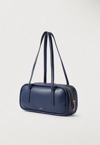 Laukkua navynsinisestä keinonahasta, jossa on kaksi olkahihnaa, suorakulmainen muoto, kultainen vetoketjusuljin ja hillitty brändilogo edessä.