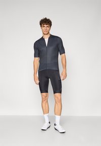 Čierny cyklistický dres s krátkymi rukávmi a predným zipsom, skombinovaný s čiernymi vystuženými šortkami. Model má na sebe biele športové topánky a ponožky.