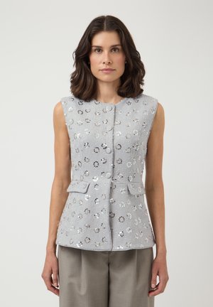 Kobieta w jasnoszarej, bezrękawnej bluzce na guziki z kwiatowymi ozdobami z cekinów oraz pasujących szarych spodniach, stojąca na jednolitym tle.