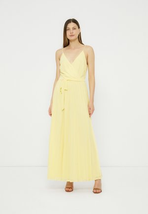 Woman standing wearing a long, pale yellow sleeveless dress with a V-neckline and waist tie, paired with brown open-toe heels.