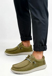Chaussures en tissu vert avec une texture lisse, bout arrondi, lacets élastiques et une semelle gris clair. Pantalons noirs retroussés visibles.